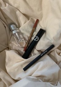 Vier cosmeticaproducten op een crèmekleurige stof: een transparante fles, twee lippotloden in bruin en perzik, en een zwarte mascara-tube.