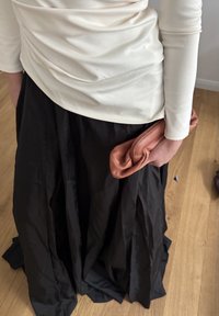 Bela dolga majica z dolgimi rokavi je spuščen nad črno, polno dolgo krilo. V eni roki drži naborano clutch torbico v rjavi barvi. Vidna je teksturirana tkanina.