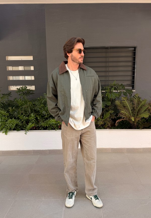 Homme portant une veste verte, une chemise blanche, un pantalon beige et des baskets, debout à l'intérieur près de plantes et d'un mur gris avec des fenêtres.