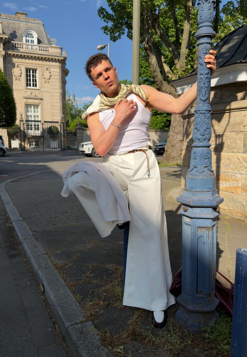 Junger Mann in weißen, weit geschnittenen Hosen und Tanktop lehnt an einem blauen Laternenpfahl und hält eine Jacke und einen Schal in der Hand auf einer sonnigen Straße in der Stadt.