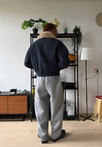 Blouson aviateur bleu marine avec un col en fausse fourrure, assorti à un pantalon de survêtement gris clair. Chaussures foncées, cadre intérieur avec des plantes et du mobilier.