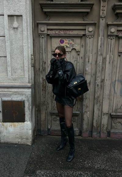 Mujer con chaqueta de cuero negra, pantalones cortos, guantes, botas, gafas de sol y un gran bolso, de pie frente a una puerta de madera envejecida con grafitis.
