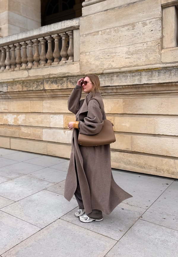 Frau mit langem braunem Mantel und Sonnenbrille trägt eine große braune Handtasche, während sie auf einem Steinpflaster neben einer beigen Steinmauer geht.