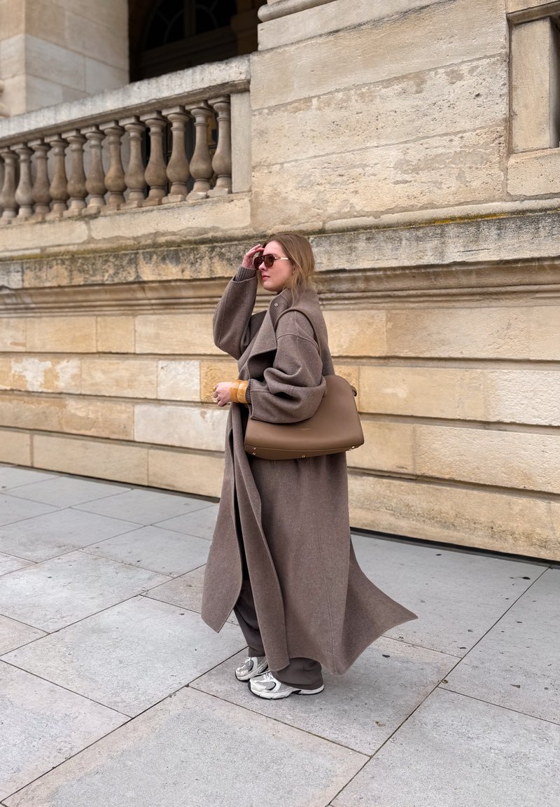 Femme portant un long manteau marron et des lunettes de soleil, tenant un grand sac à main marron tout en marchant sur un trottoir en pierre près d'un mur en pierre beige.