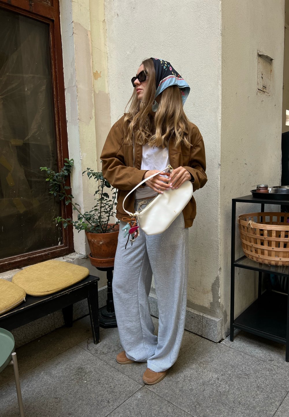 Mulher com jaqueta castanha, camisa branca, calças risca de giz e lenço na cabeça segura uma bolsa branca, em pé dentro de casa perto de uma planta em vaso e um banco com almofadas.