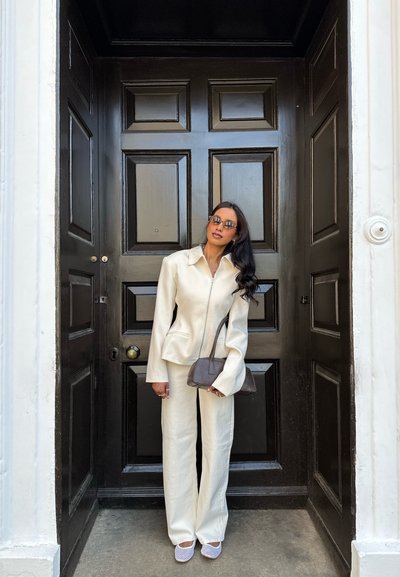 Mujer de pie frente a una gran puerta de madera oscura, vestida con un traje pantalón color crema, gafas de sol, sosteniendo un bolso marrón y mirando hacia adelante.