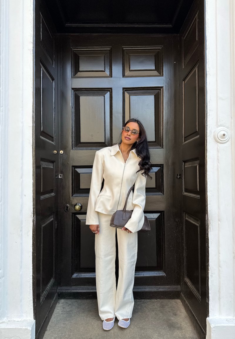 Femme debout devant une grande porte en bois foncé, vêtue d'un tailleur-pantalon crème, lunettes de soleil, tenant un sac à main brun et faisant face à l'avant.