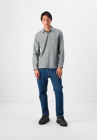 Camicia grigia con bottoni e tasca, con un logo verde. Abbinata a jeans blu e sneakers nere con suola testurizzata.