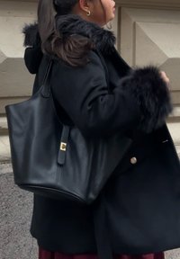 Sac cabas en cuir noir avec une finition texturée, doté d'une fermeture à clapet dorée et d'une silhouette large et épurée. Mis en valeur par un manteau en laine.