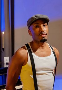 Homem com uma barba cuidadosamente aparada, a usar um boné cinza e uma camisola sem mangas branca, complementada por suspensórios pretos. O fundo apresenta uma iluminação suave.