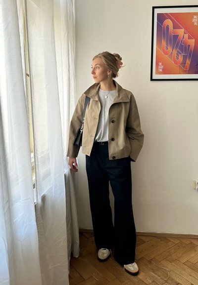 Chaqueta cropped beige con grandes botones sobre una blusa gris, combinada con pantalones de pierna ancha oscuros y zapatillas color crema, de pie junto a una ventana.