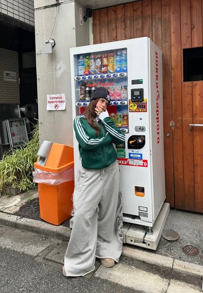 Máquina expendedora con cuerpo blanco; etiquetas de bebidas brillantes visibles. Cubo de basura naranja cerca, con un fondo de pared de madera y elementos de concreto.