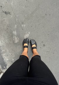 Bailarinas negras con punta redondeada, que cuentan con un pequeño lazo decorativo y correas elásticas. Llevadas con leggings negros sobre una acera gris.
