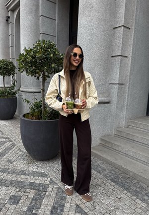Mujer con el pelo largo que lleva gafas de sol y chaqueta color crema, sosteniendo dos bebidas para llevar, de pie junto a escalones de piedra y grandes plantas en macetas.