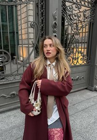 Manteau oversize bordeaux, chemise blanche, cravate beige, collants à motifs. Petit sac tissé avec détails décoratifs. Portail en fer forgé complexe en arrière-plan.