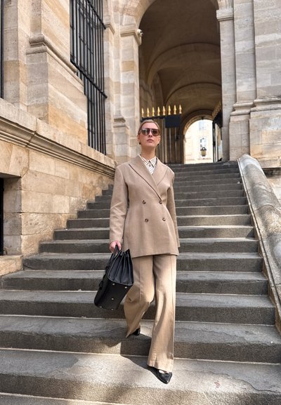 Mujer con traje beige y gafas de sol bajando unas escaleras de piedra frente a un edificio histórico, llevando un bolso negro.