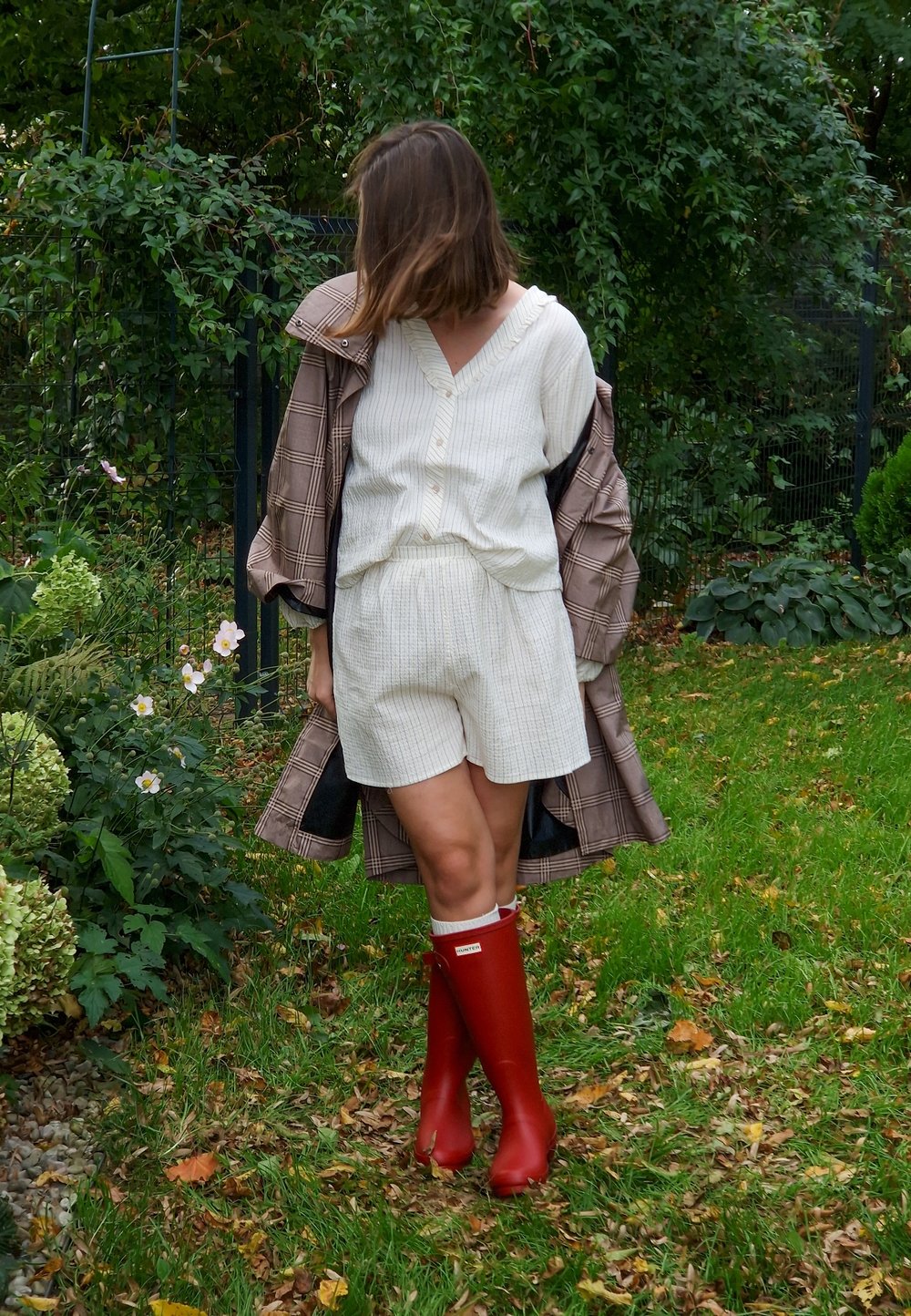 Cortos de rayas beige y blancas con una blusa a juego, combinados con un abrigo de cuadros marrón y botas de goma rojas. Vegetación verde y flores de fondo.
