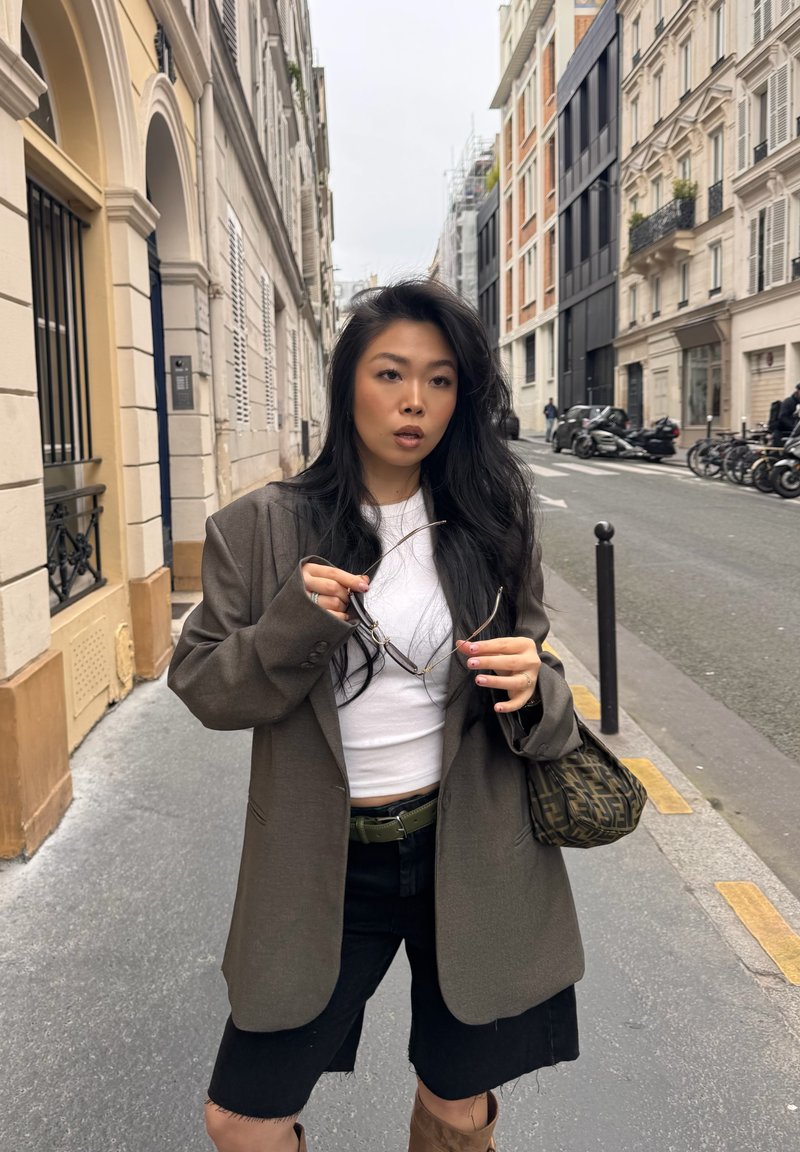 Femme aux longs cheveux noirs tenant des lunettes de soleil dans une rue de la ville, portant un blazer marron, une chemise blanche, un short noir et portant un sac à bandoulière à motifs.