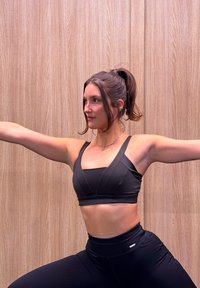 Femme avec queue de cheval en tenue de sport noire étirant les bras en posture de yoga debout devant un mur en bois.