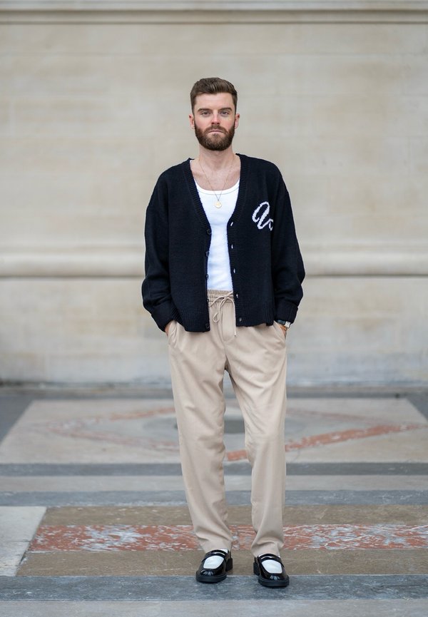 Cardigan preto de malha com detalhes em letras brancas, top de alças branco, calças bege e sapatos pretos com detalhes brancos, de pé sobre um chão estampado.
