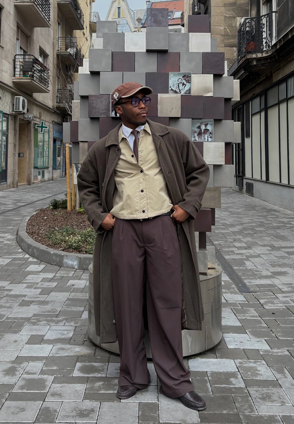 Καφέ μάλλινο παλτό, μπεζ πουκάμισο με κουμπιά, καφέ γραβάτα και φαρδιές καφέ παντελόνες. Αξεσουάρ περιλαμβάνουν γυαλιά και καφέ καπέλο. Υφασμάτινο φόντο.