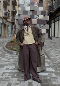 Καφέ μάλλινο παλτό, μπεζ πουκάμισο με κουμπιά, καφέ γραβάτα και φαρδιές καφέ παντελόνες. Αξεσουάρ περιλαμβάνουν γυαλιά και καφέ καπέλο. Υφασμάτινο φόντο.