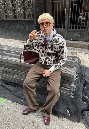Chaqueta con estampado de vaca multicolor y cremallera, blanca con manchas negras, combinada con pantalones de pierna ancha marrones. Gafas de sol naranjas en forma circular y bolso burdeos.