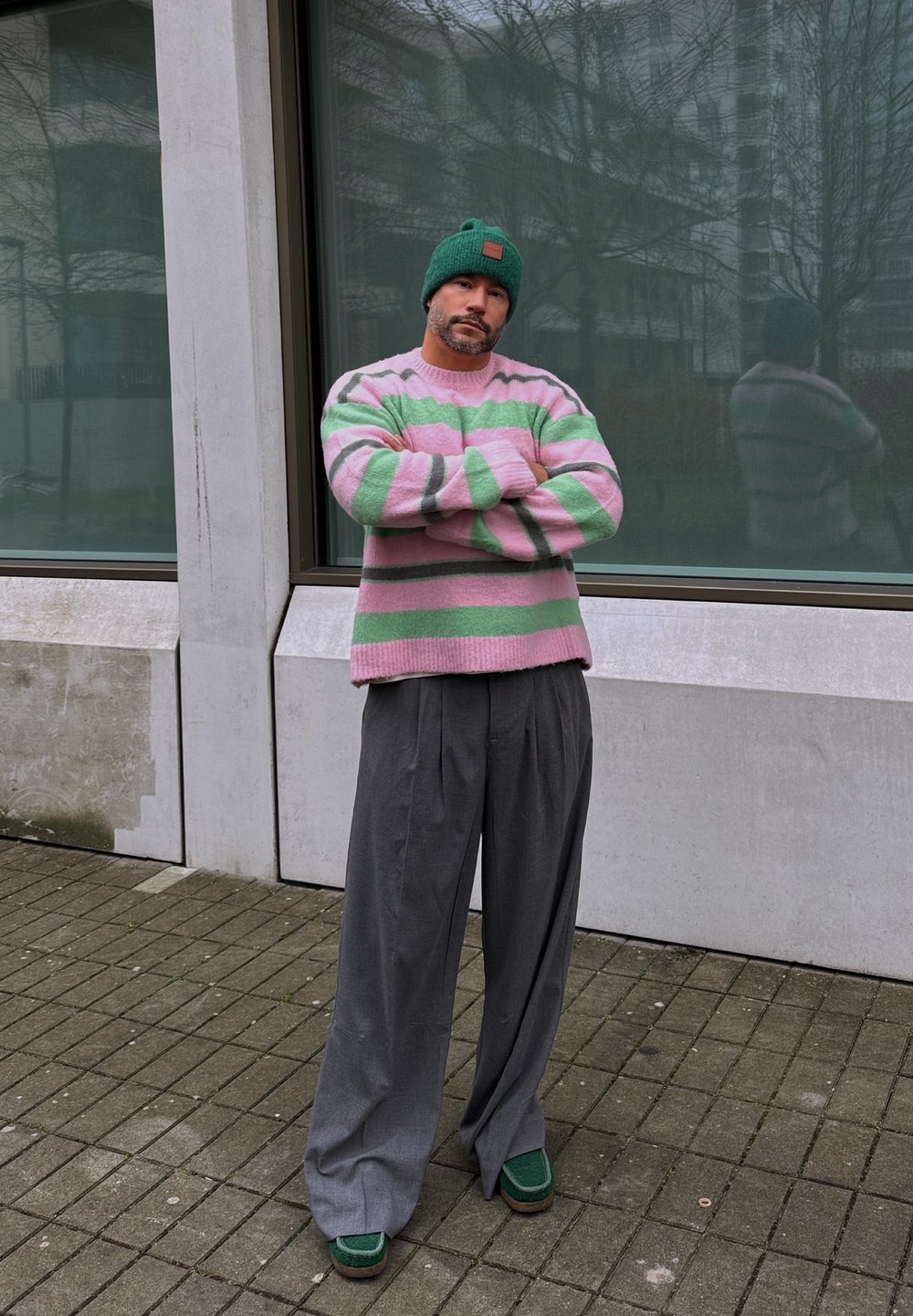 Suéter a rayas rosa y verde, pantalones grises de pierna ancha y gorro verde. El atuendo tiene un ajuste holgado y texturas contrastantes.