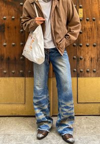 Baggy, lyseblå denimjeans med falmede mønstre, kombinert med en lett brun jakke og en hvit totebag. Teksturert cowboystøvler har dekorative detaljer.