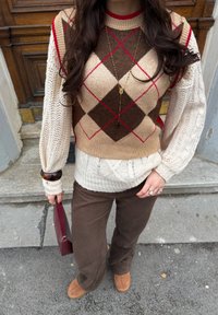 Pull en tricot présentant un motif argyle en marron, rouge et beige avec des manches en tricot torsadé. Associé à un pantalon marron et des chaussures en daim.