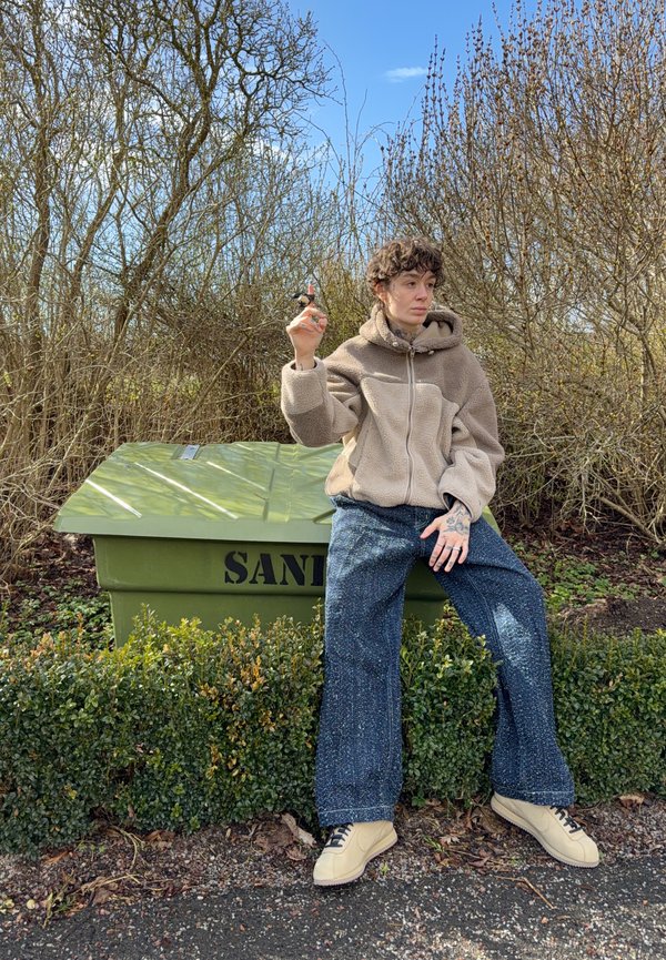 Personne aux cheveux bouclés portant une veste beige et un jean large, assise sur un conteneur à sable vert, tenant un objet, entourée de buissons sans feuilles.