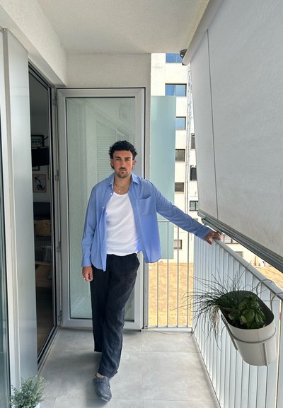 Camisa de manga larga azul claro sobre una camiseta blanca y pantalones negros, de pie en un balcón con puertas de cristal y plantas en macetas.