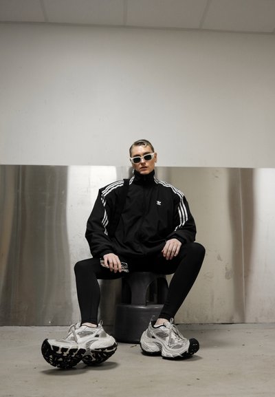 Chaqueta de chándal negra de Adidas con rayas blancas, leggings negros y zapatillas gruesas grises. Los accesorios incluyen gafas de sol blancas y una funda de teléfono transparente.