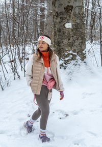 Femme portant des vêtements d'hiver et un bandeau marchant sur un sentier forestier enneigé près d'un arbre avec un panneau vert de premiers secours.