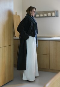 Črni velikanski plašč, siva šal in bele hlače širokih hlačnic z gladkim teksturo. Minimalistična kuhinja v ozadju z lesenimi elementi.