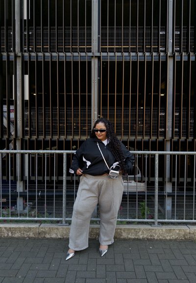 Chaqueta deportiva negra con rayas blancas, pantalones de chándal grises y tacones puntiagudos plateados. Cabello largo y rizado y gafas de sol elegantes. Fondo urbano.
