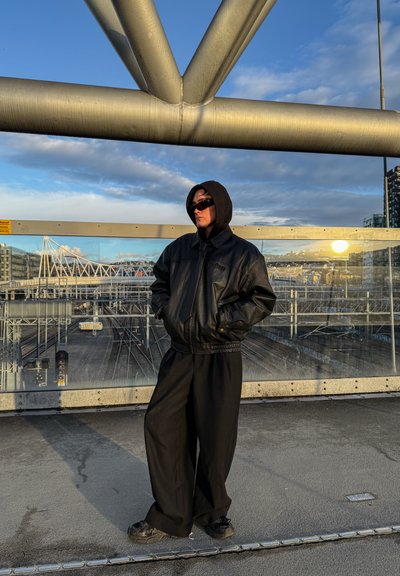 Chaqueta bombér negra de cuero con capucha, pantalones negros holgados y gafas de sol. De pie contra una estructura metálica con vías de tren al fondo.
