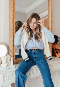 Uma modelo usa uma camisa de botão listrada azul clara, atada à cintura, combinada com umas calças de ganga largas azuis e um cardigan creme com detalhes verdes.