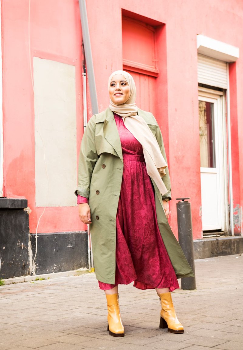 Green trench coat over a burgundy dress, featuring floral texture. Beige scarf, yellow ankle boots. City setting with pink wall backdrop.