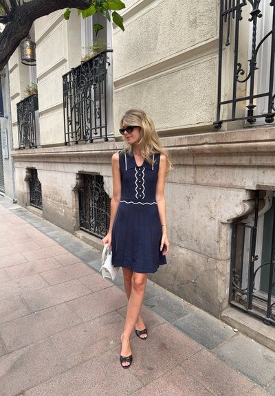 Vestido sin mangas azul marino con ribetes blancos y botones decorativos; falda plisada. Usado con sandalias negras y sosteniendo un bolso blanco.