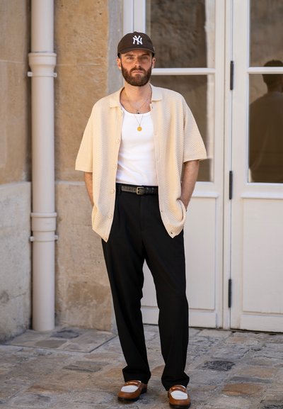 Camisa de manga corta texturizada beige sobre una camiseta blanca, combinada con pantalones negros y zapatos marrón y blancos. Los accesorios incluyen una gorra negra y collares.