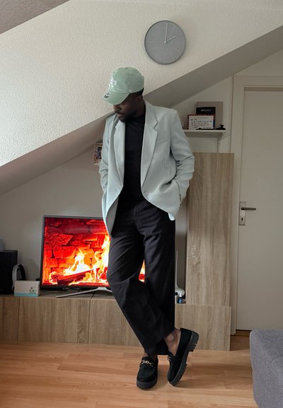 Blazer azul claro sobre una camisa negra, combinado con pantalones negros y zapatos verdes. El fondo presenta un televisor que muestra un efecto de fuego.