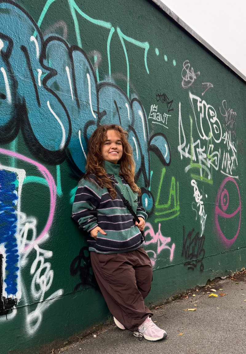 Una persona si appoggia a un muro coperto di graffiti verdi, indossando una giacca di pile a righe verdi, pantaloni larghi marroni e sneakers rosa chiaro.