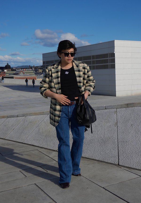 Persona con gafas de sol que lleva una chaqueta de cuadros, camisa negra y vaqueros caminando al aire libre cerca de edificios modernos bajo un cielo azul claro.