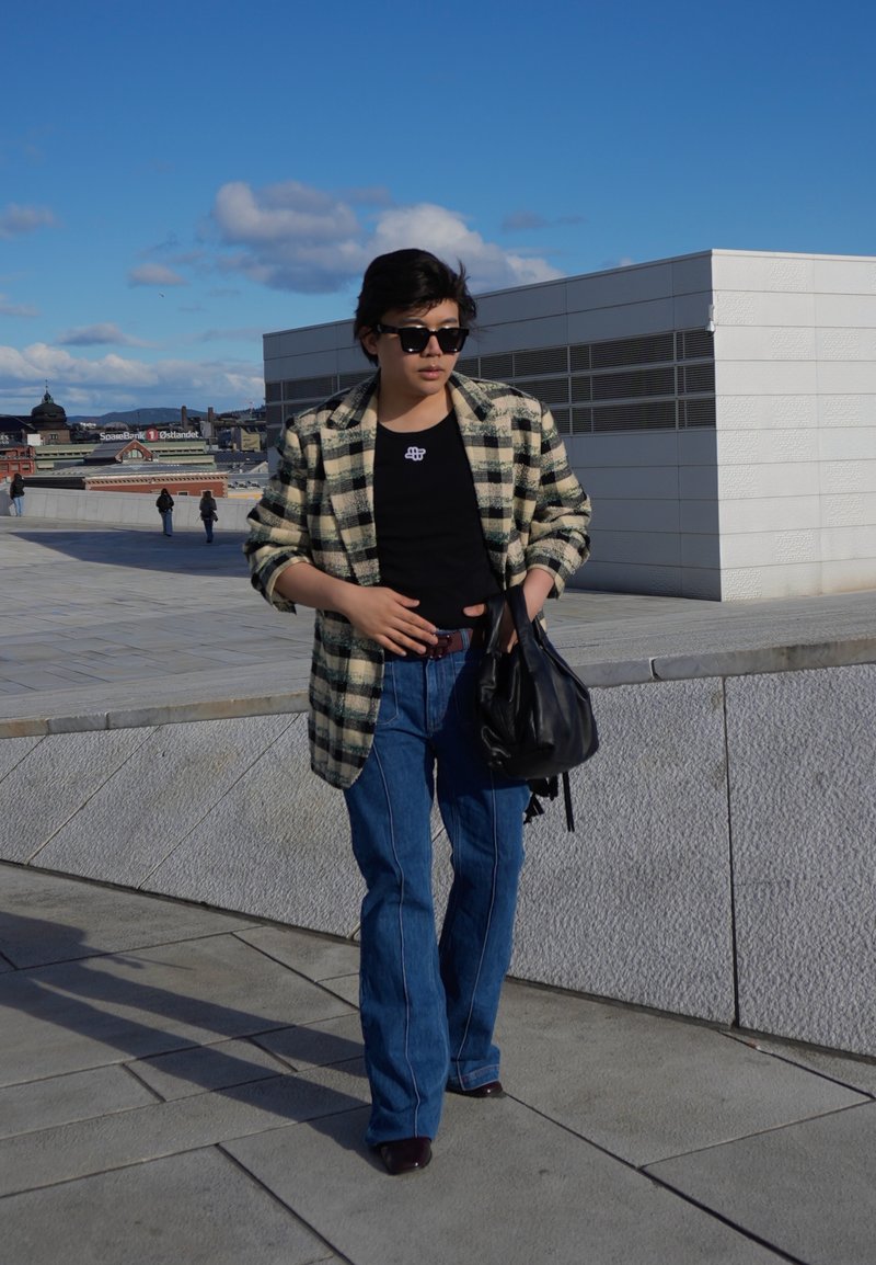 Personne portant des lunettes de soleil, une veste à carreaux, une chemise noire et un jean, marchant à l'extérieur près de bâtiments modernes sous un ciel bleu clair.