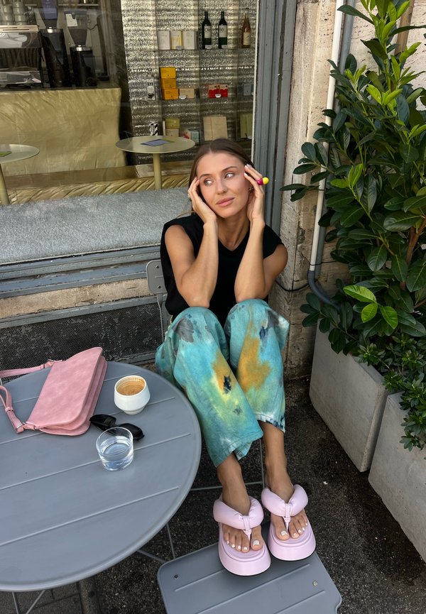 Frau sitzt draußen vor einem Café, trägt bunte Hosen und pinke Plateau-Sandalen, mit einer pinken Tasche, Kaffee und einem Glas Wasser auf dem Tisch.