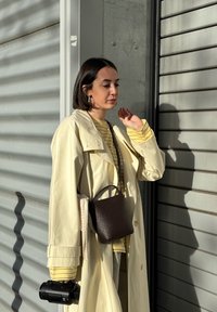 Light yellow trench coat over a striped top, holding a small brown textured bag and a camera, with a shadow cast on a grey backdrop.