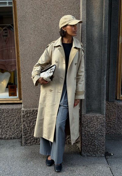 Gabardina beige con diseño de doble botonadura, pantalones grises anchos, top negro y una gorra beige. Sosteniendo un clutch estampado, de pie frente a una pared texturizada.