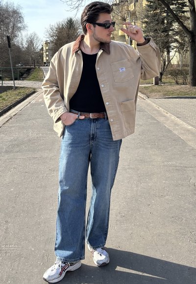 Chaqueta de algodón beige con un cuello marrón, camisa negra, vaqueros azules de corte holgado y zapatillas blancas con acentos en rojo y negro. Entorno al aire libre.
