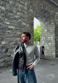 Camisa henley de color gris claro, chaqueta de cuero oscura sobre el hombro, jeans de denim azul; de pie bajo un arco de piedra con árboles verdes de fondo.